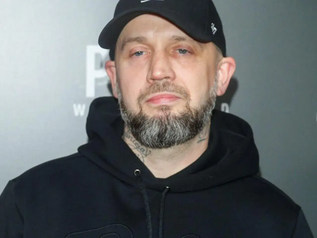 Mężczyzna w czarnej bluzie z kapturem i czapce z daszkiem, z brodą i tatuażami na szyi. Pikers ile ma lat?
