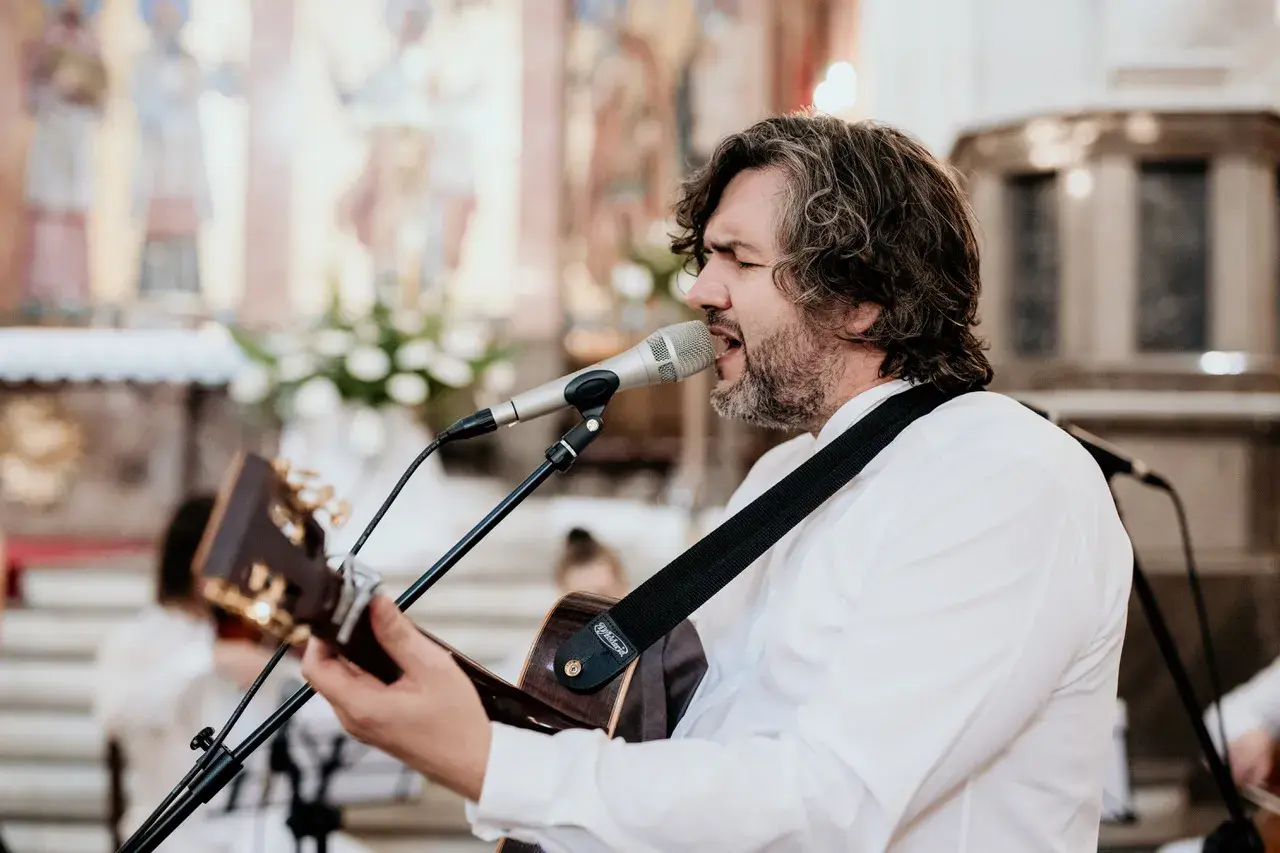 Mężczyzna z gitarą śpiewa pieśni chrześcijańskie w kościele. W tle widać ołtarz i obrazy.