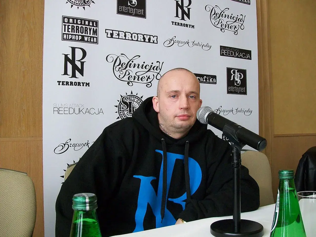 Peja w czarnej bluzie z niebieskim logo, przed mikrofonem. Na ścianie za nim napisy: "TERRORYM", "REEDUKACJA", "Definicja Peja". Wiecie co z nim zrobić.
