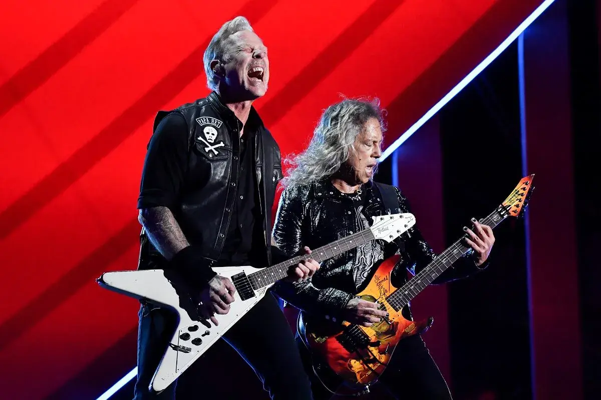 Dwóch gitarzystów zespołu Metallica gra na scenie na tle czerwonej kurtyny.
