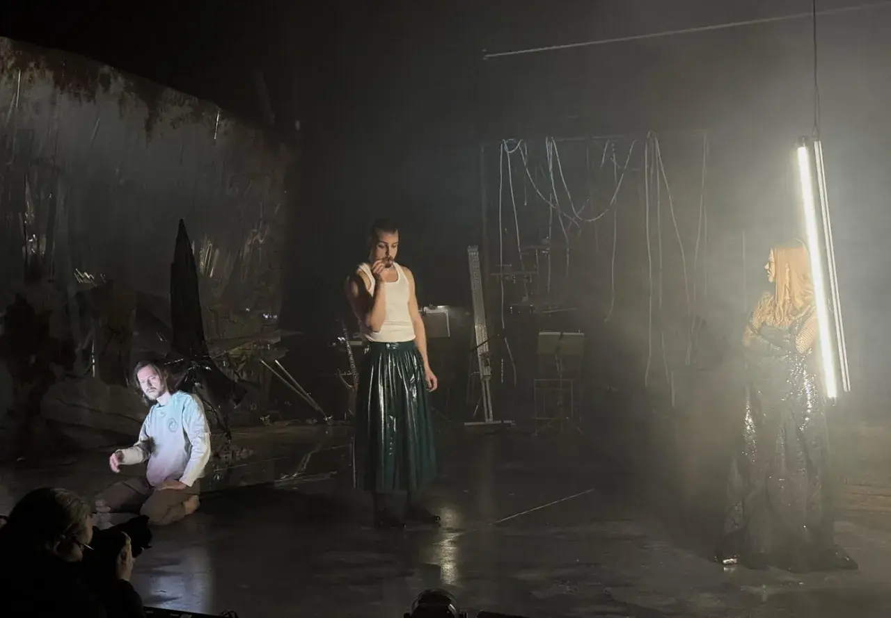 Trzej aktorzy na scenie, jeden klęczy, drugi stoi w błyszczącej spódnicy, trzecia w połyskującej sukni. Scenografia sugeruje **epic the musical**.