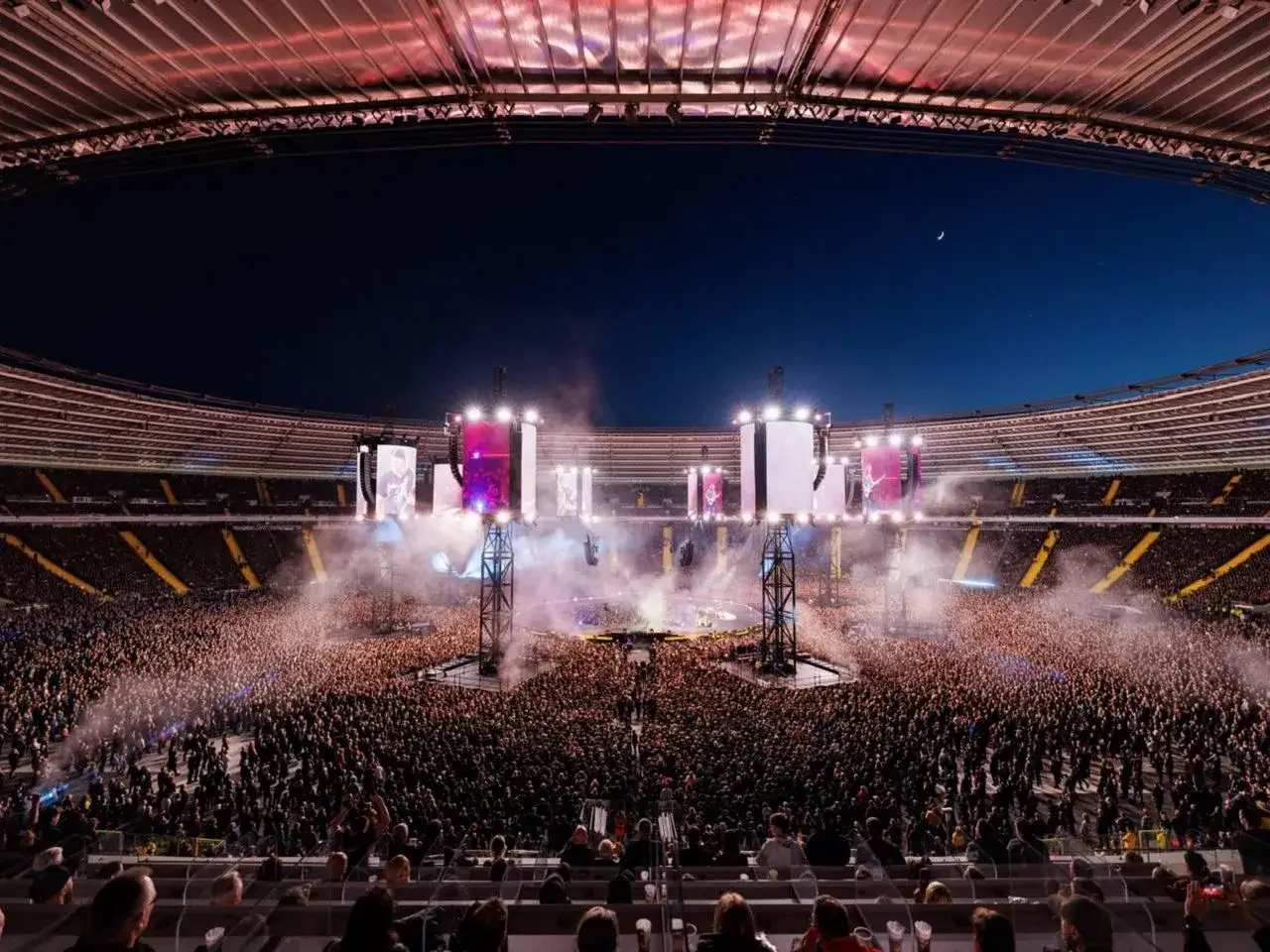 Ogromny tłum na koncercie w stadium, scena oświetlona, dym i światła tworzą atmosferę, która sprawia, że wszystko wydaje się niemal blackened.