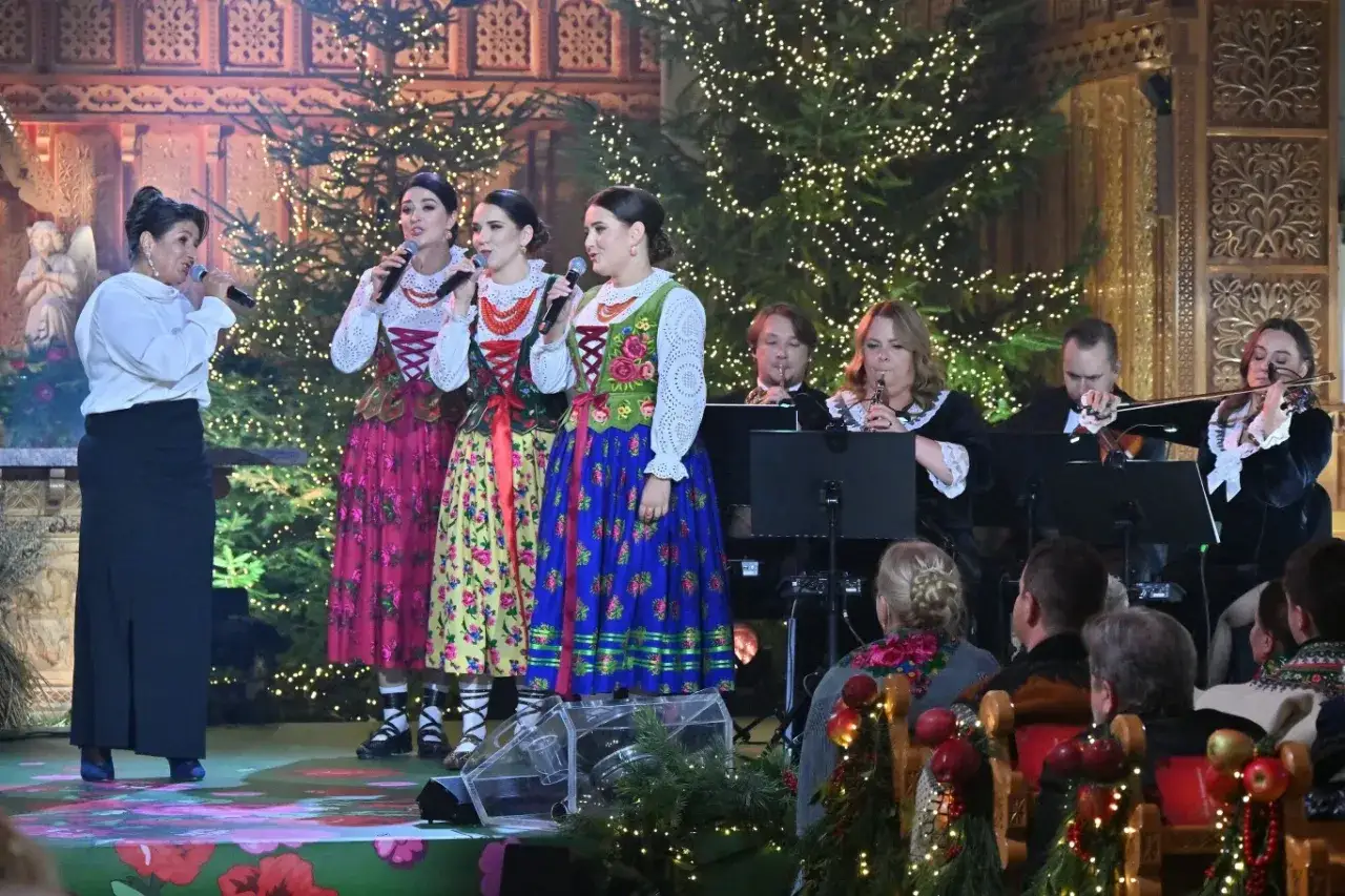 Zespół w strojach ludowych śpiewa kolędy przy akompaniamencie orkiestry. W tle choinki i świąteczne dekoracje.
