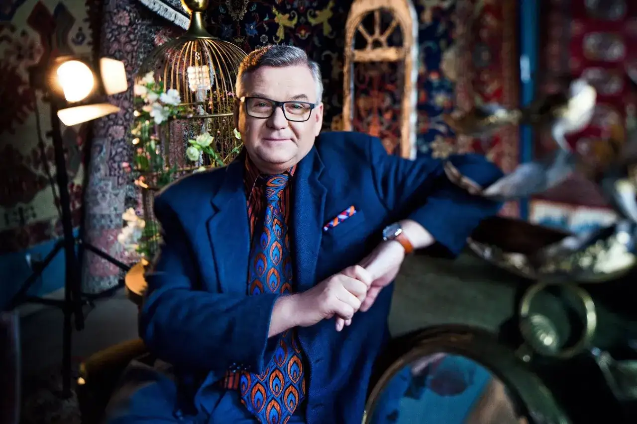 Artur Andrus w eleganckim garniturze, z kolorowym krawatem, siedzi w otoczeniu dekoracji. Jego piosenki artura andrusa alfabetycznie to bogactwo słów i melodii.