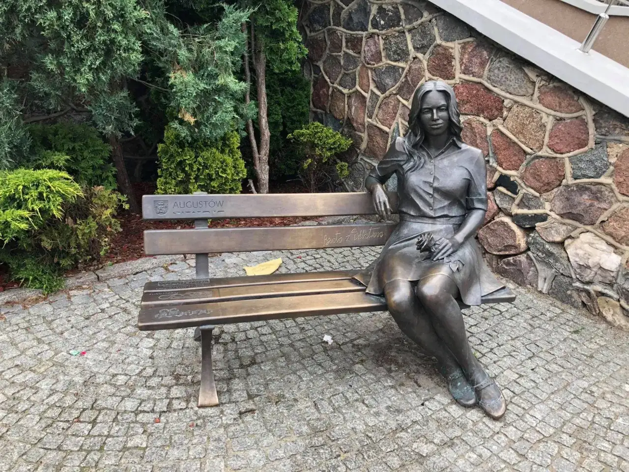 Ławeczka z rzeźbą kobiety, która trzyma różę. Na ławce widnieje napis