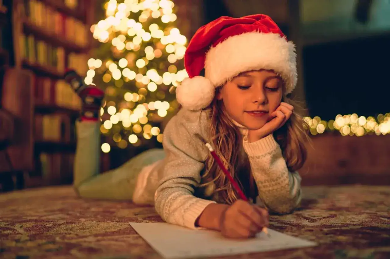 Dziewczynka w czapce Mikołaja pisze list do Mikołaja przy choince.