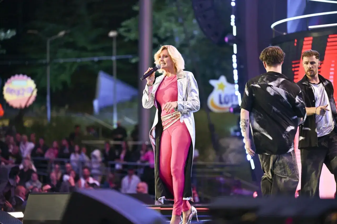 Helena Vondráčková w różowym stroju i srebrnym płaszczu śpiewa na scenie, obok niej dwaj mężczyźni.