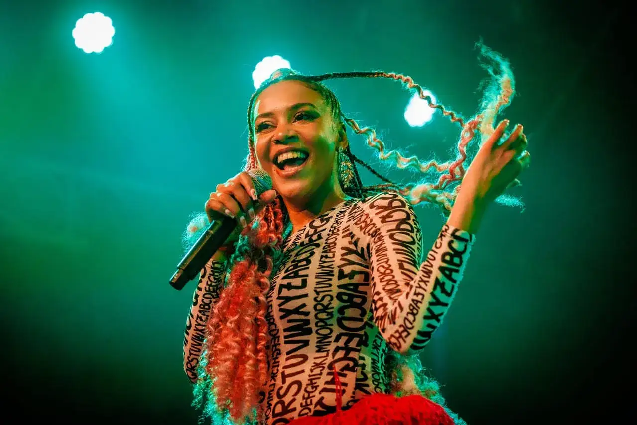 Uśmiechnięta artystka z kolorowymi warkoczami śpiewa do mikrofonu. Występuje na scenie, jak wielu hiszpańskich piosenkarzy.