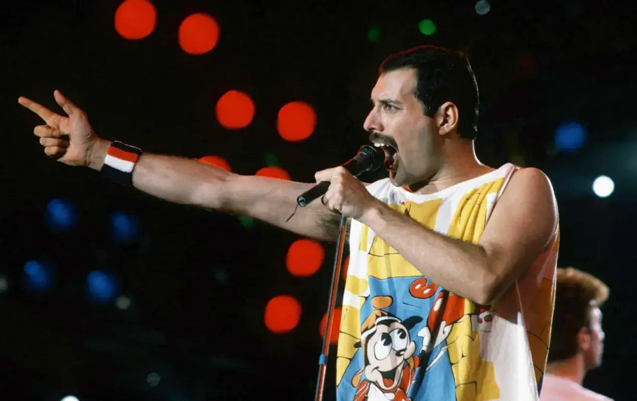 Freddie Mercury śpiewa na koncercie, wskazując palcem. Jego koszulka z Goofym przypomina, że