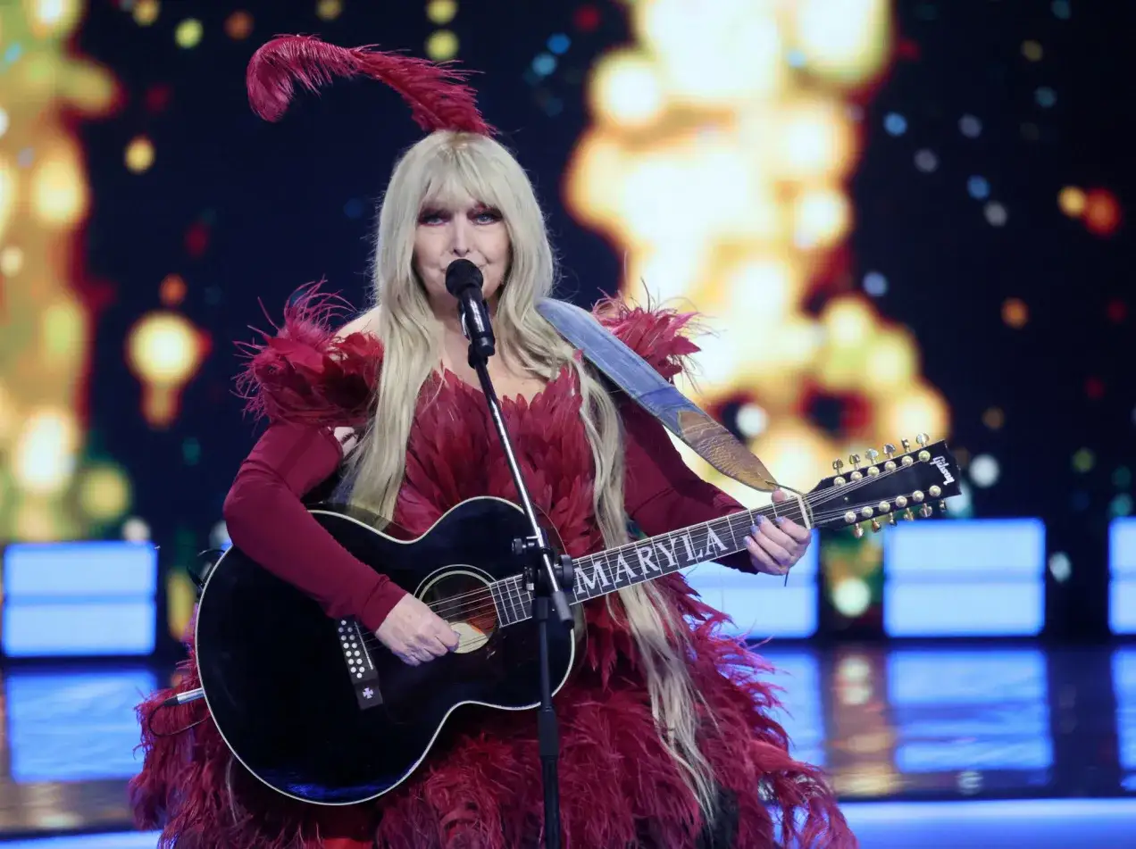 Maryla Rodowicz w piórach i z gitarą, gotowa do wykonania swoich piosenek alfabetycznie.