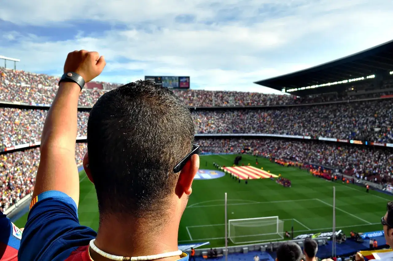 Widok z trybun stadionu na mecz, kibic z uniesioną pięścią, słychać hymn Barcelony.