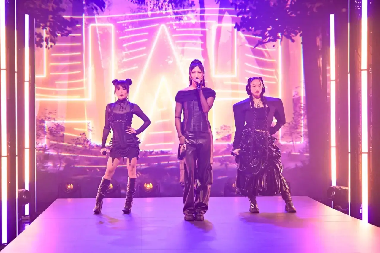 Trzy artystki k-pop prezentują swoje utwory na scenie, w neonowym świetle i futurystycznych strojach.