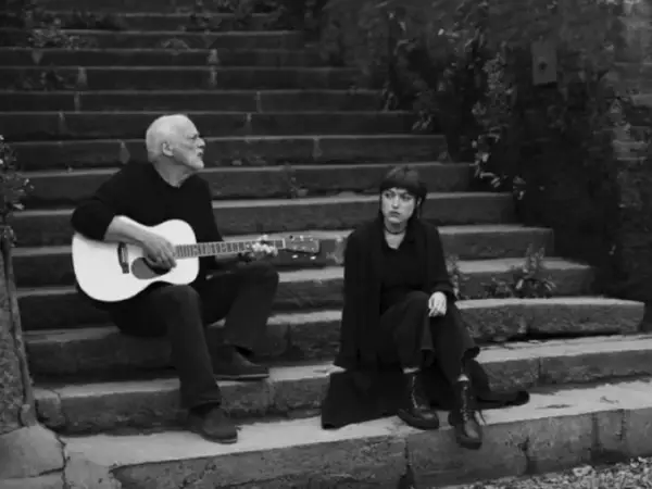 David Gilmour gra na gitarze, a obok niego siedzi jego córka.